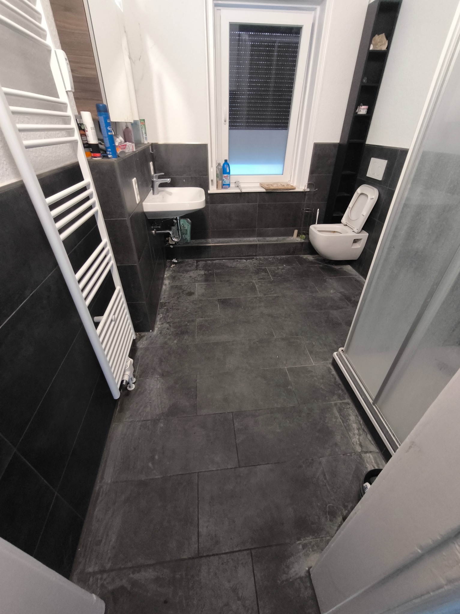 Ciemna łazienka z umywalką, toaletą i grzejnikiem drabinkowym. Widoczne ślady użytkowania na podłodze i ścianach. Potencjalne sprzątanie po remoncie lub gruntowne czyszczenie.