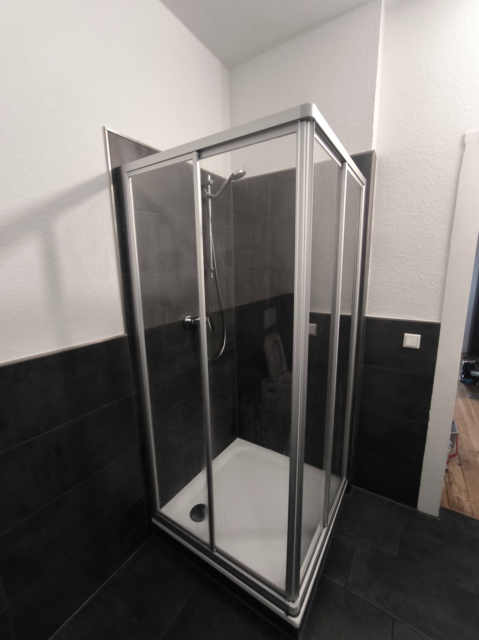 Nowoczesna kabina prysznicowa z przesuwnymi drzwiami, wykończona czarnymi płytkami i białymi ścianami. Widoczny brodzik i bateria prysznicowa.