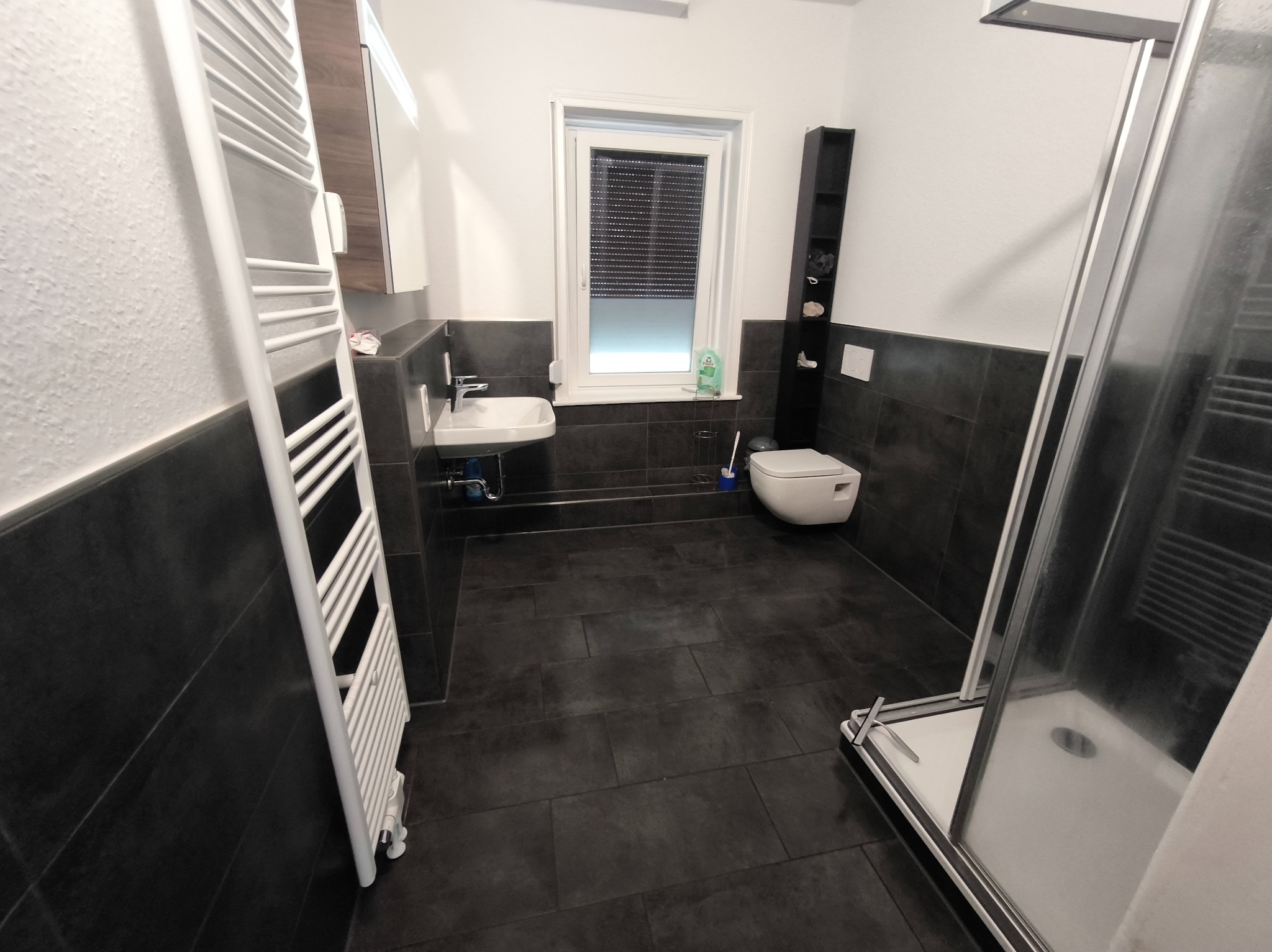 Nowoczesna łazienka z czarnymi płytkami, białym wyposażeniem i grzejnikiem drabinkowym. Widoczna kabina prysznicowa, umywalka i toaleta. Minimalistyczny design.