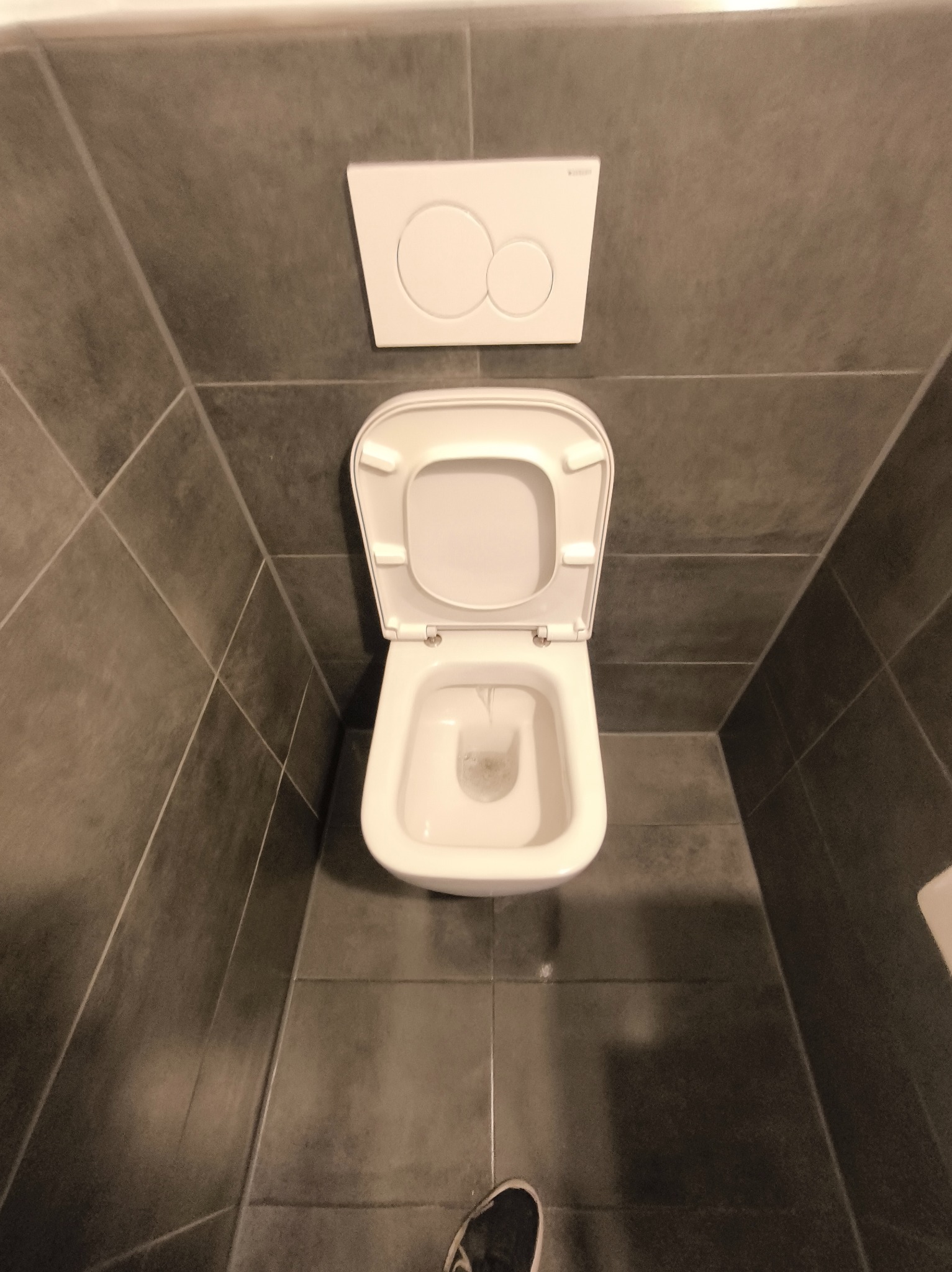 Biała toaleta z otwartą klapą na tle szarych płytek. Widoczny przycisk spłukiwania i fragment buta w dolnej części kadru. Ujęcie z góry.