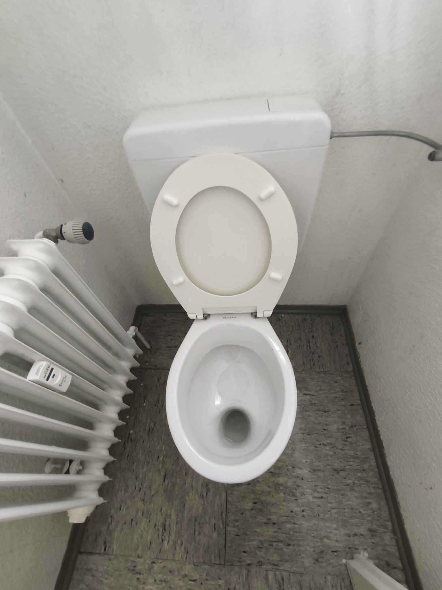 Biała toaleta z otwartą klapą, widok z góry, obok grzejnik. Szara podłoga i białe ściany. Ujęcie z bliska, oświetlenie naturalne.