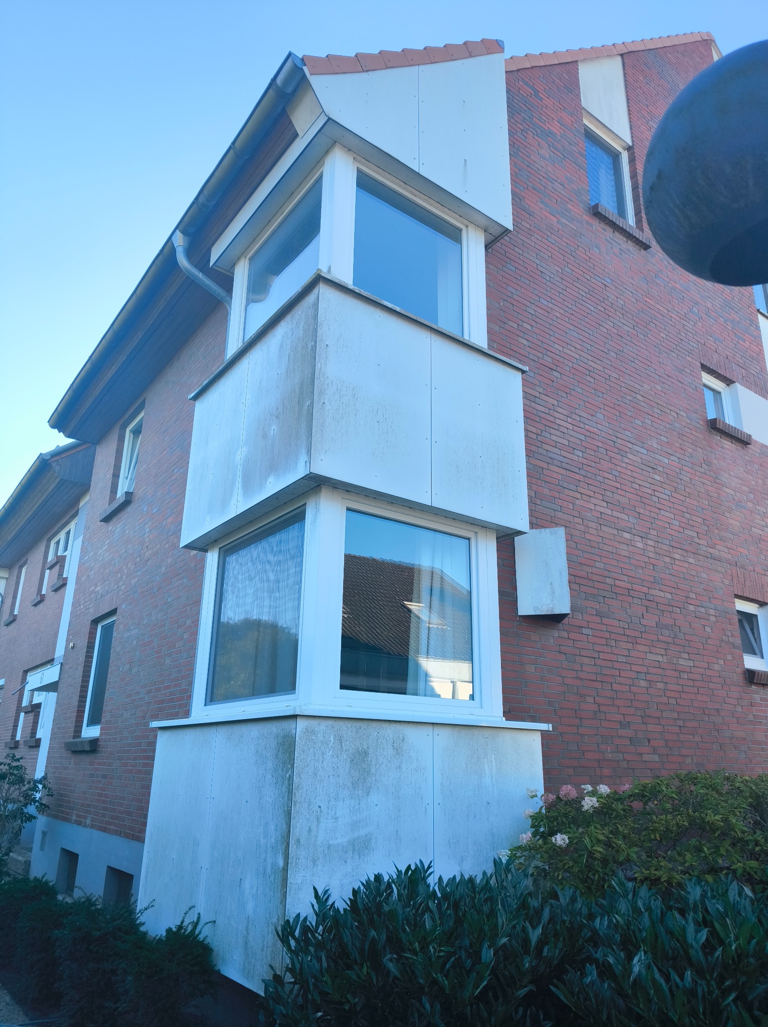 Dwukondygnacyjny wykusz z białymi ramami okiennymi na tle ceglanej ściany budynku, widoczny z dołu pod kątem. Na dole krzewy.