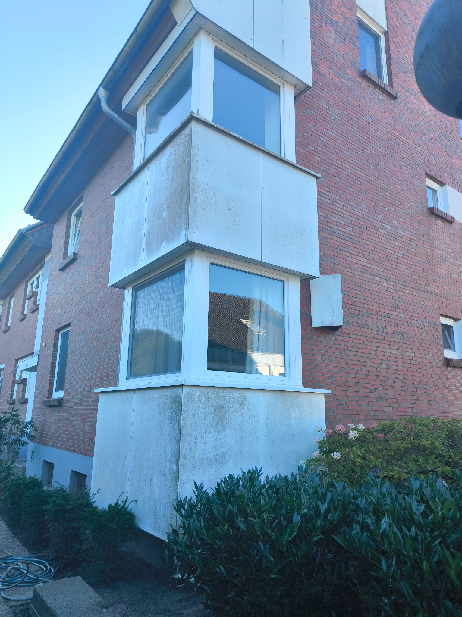 Zewnętrzny narożnik budynku z cegły z wystającym, zabrudzonym balkonem z dwoma oknami. Widoczne krzewy i fragment węża ogrodowego.