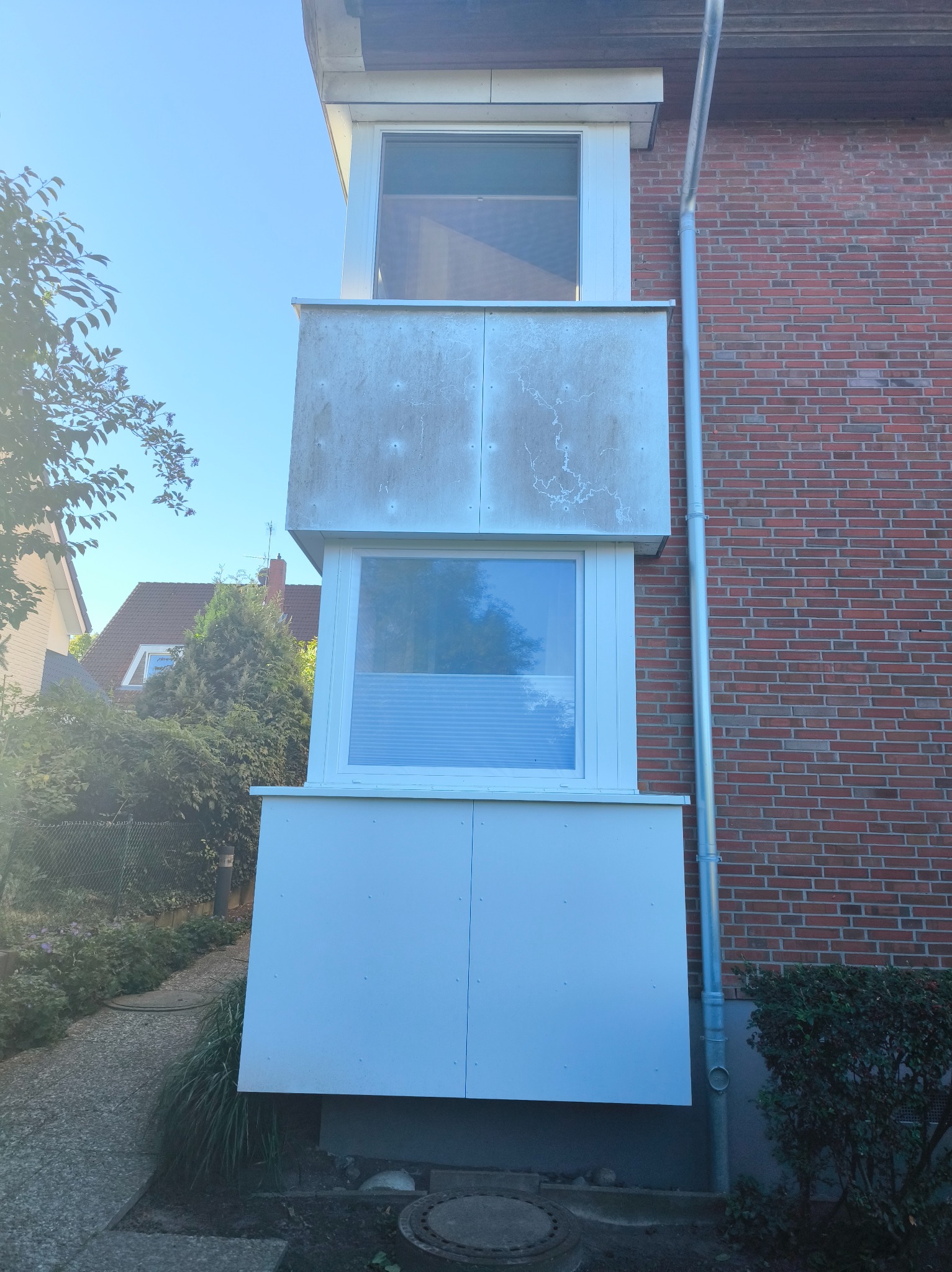 Zewnętrzny balkon z dwoma oknami, z widocznymi śladami zabrudzeń na elewacji, przylegający do ceglanego budynku z rynną. Widok z dołu na balkon.