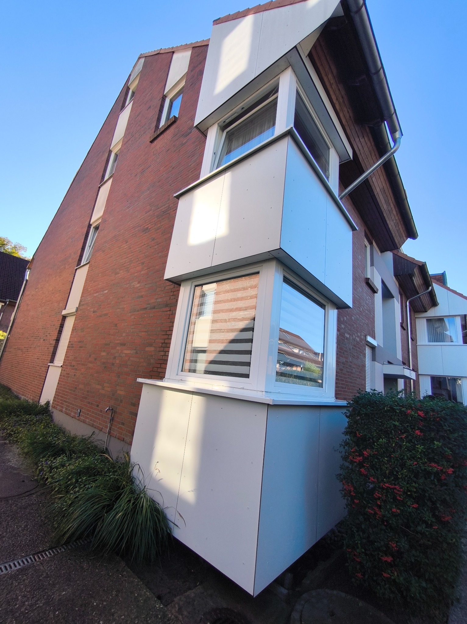 Ceglany budynek z białym, wystającym oknem balkonowym i roletami, widziany z dołu pod kątem, z zielenią u podstawy i błękitnym niebem w tle.