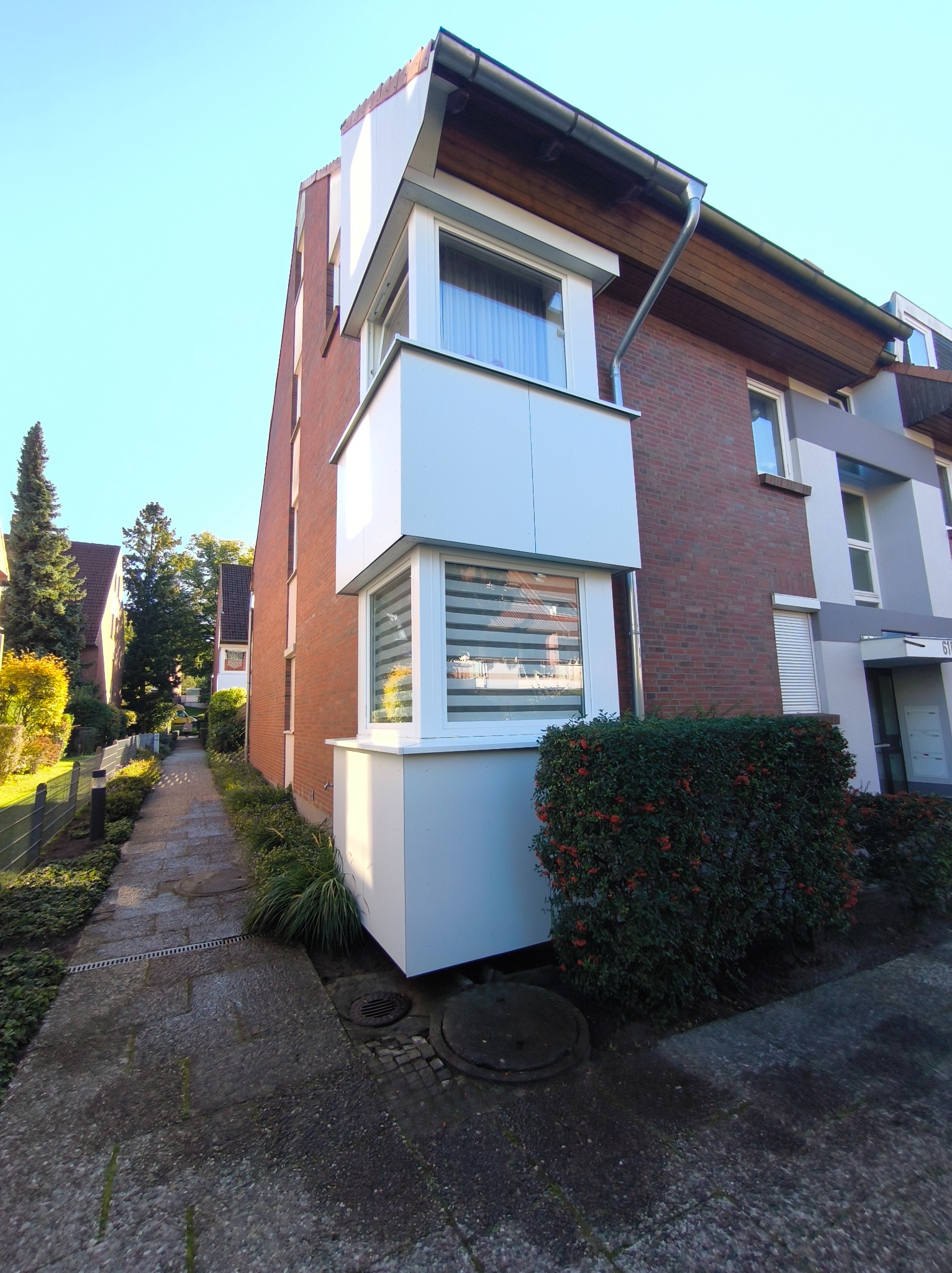 Widok z dołu na dwupiętrowy budynek z cegły z białym, wystającym balkonem i oknami z żaluzjami. Obok budynku rośnie krzew z czerwonymi owocami.