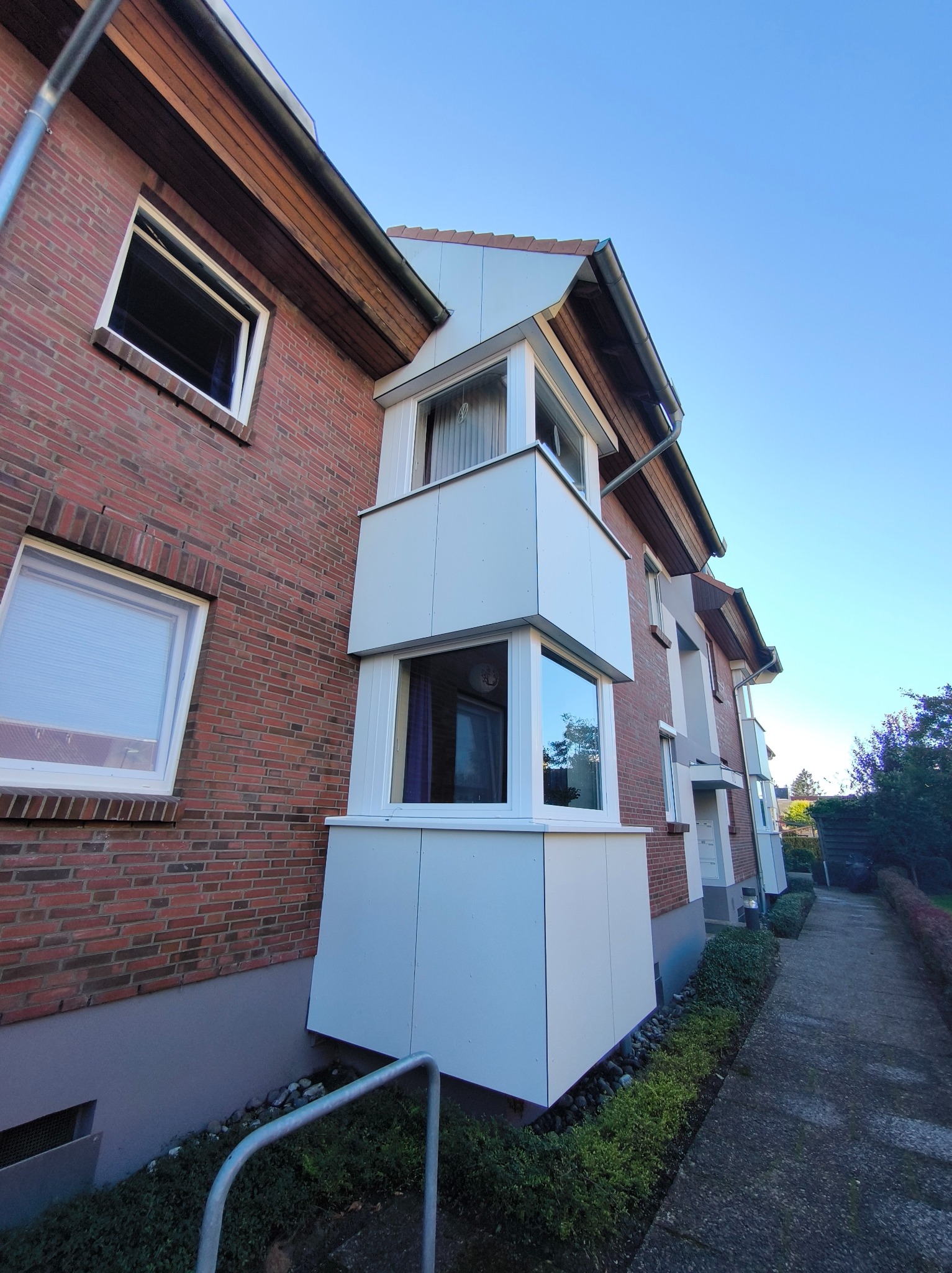 Ceglany budynek z białymi balkonami, widok z dołu, błękitne niebo. Balkony z białymi panelami, okna z białymi ramami. Ścieżka obok budynku.