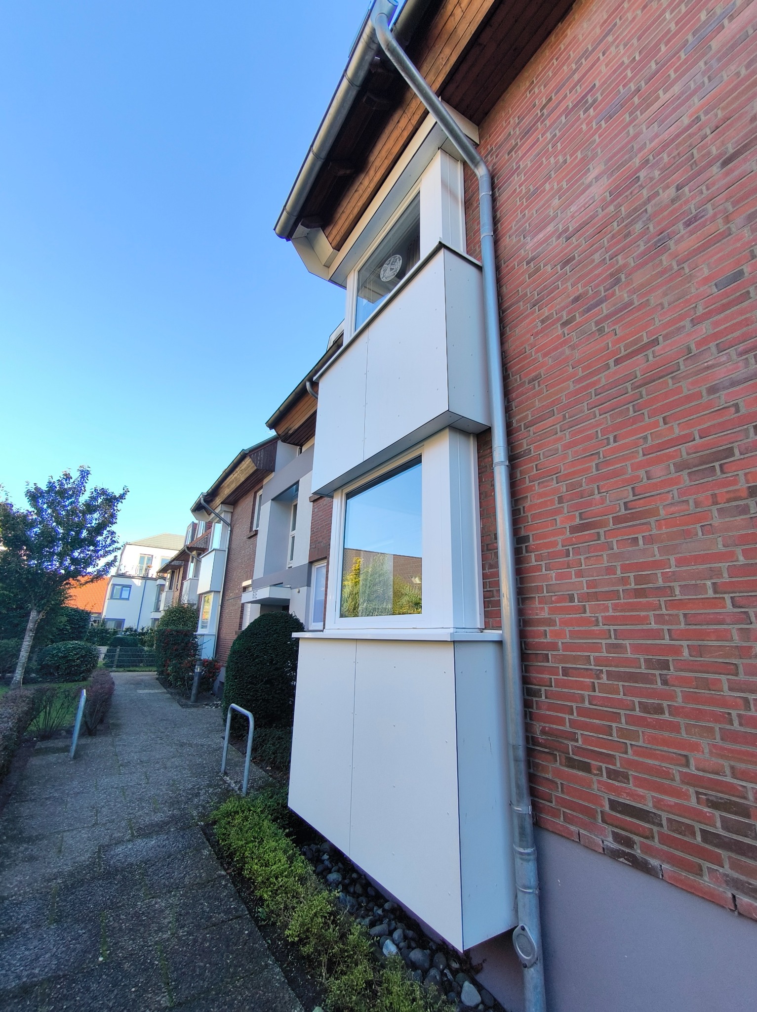 Fasada budynku z cegły z białymi elementami balkonu i rynnami, widok z dołu w słoneczny dzień. Perspektywa ulicy z zielenią.