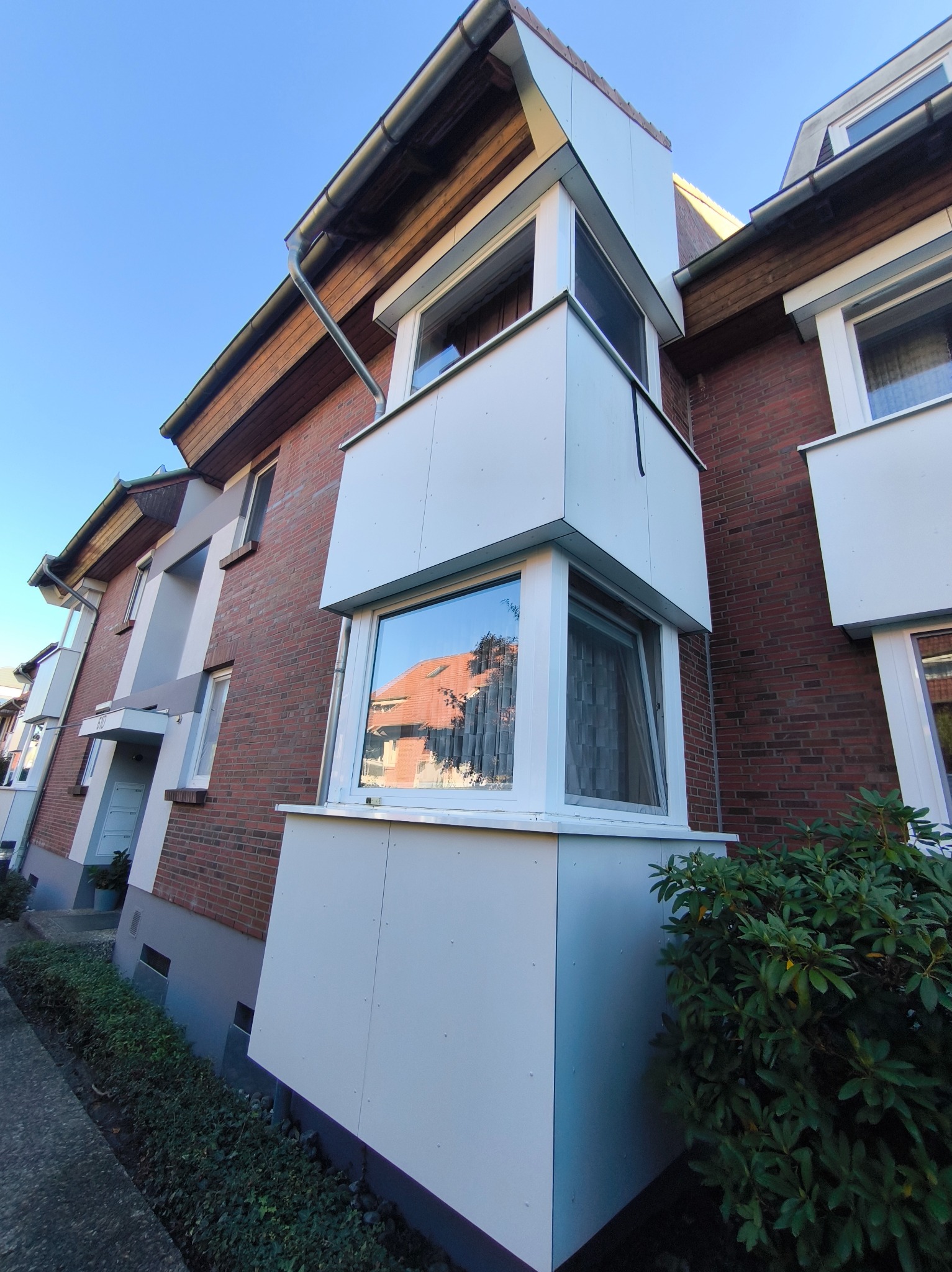 Nietypowy, kanciasty balkon z białymi panelami na tle ceglanej elewacji budynku, widok z dołu, błękitne niebo, krzew przy chodniku.