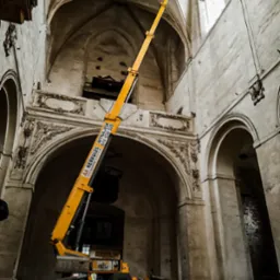 Żółty podnośnik koszowy wewnątrz zabytkowego kościoła z dekoracyjnymi łukami i filarami, platforma podnośnika przy oknie.