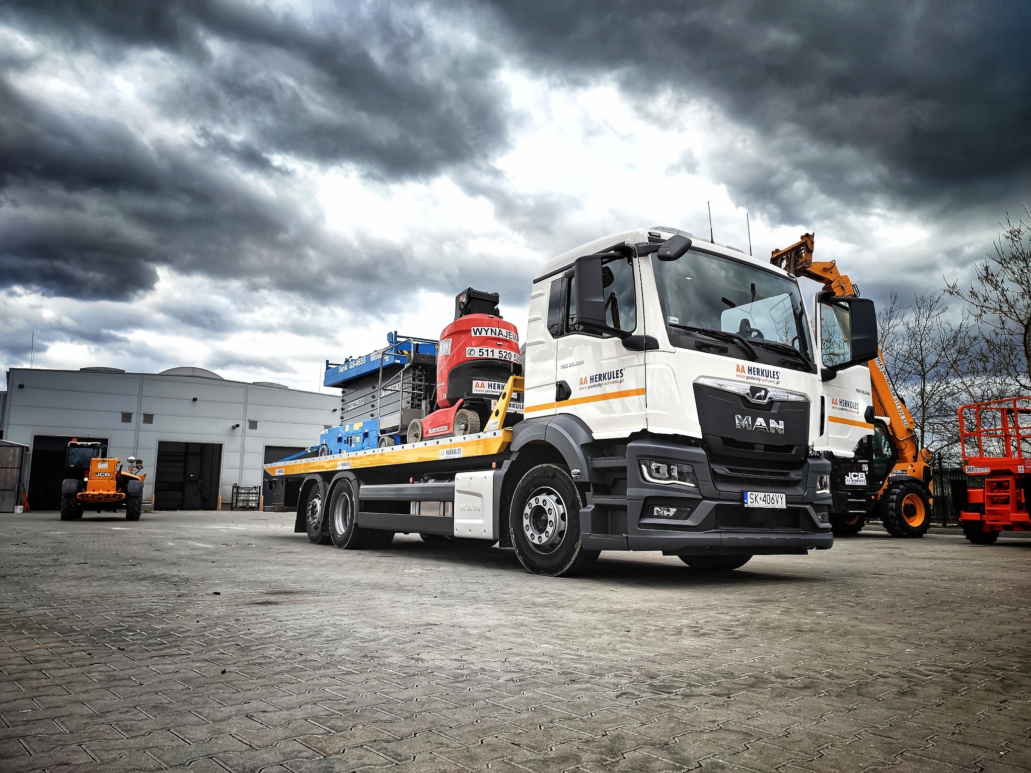 Ciężarówka MAN z platformą załadunkową przewożąca podnośnik koszowy na tle pochmurnego nieba, w tle widoczny pomarańczowy podnośnik teleskopowy JCB i budynek magazynowy.