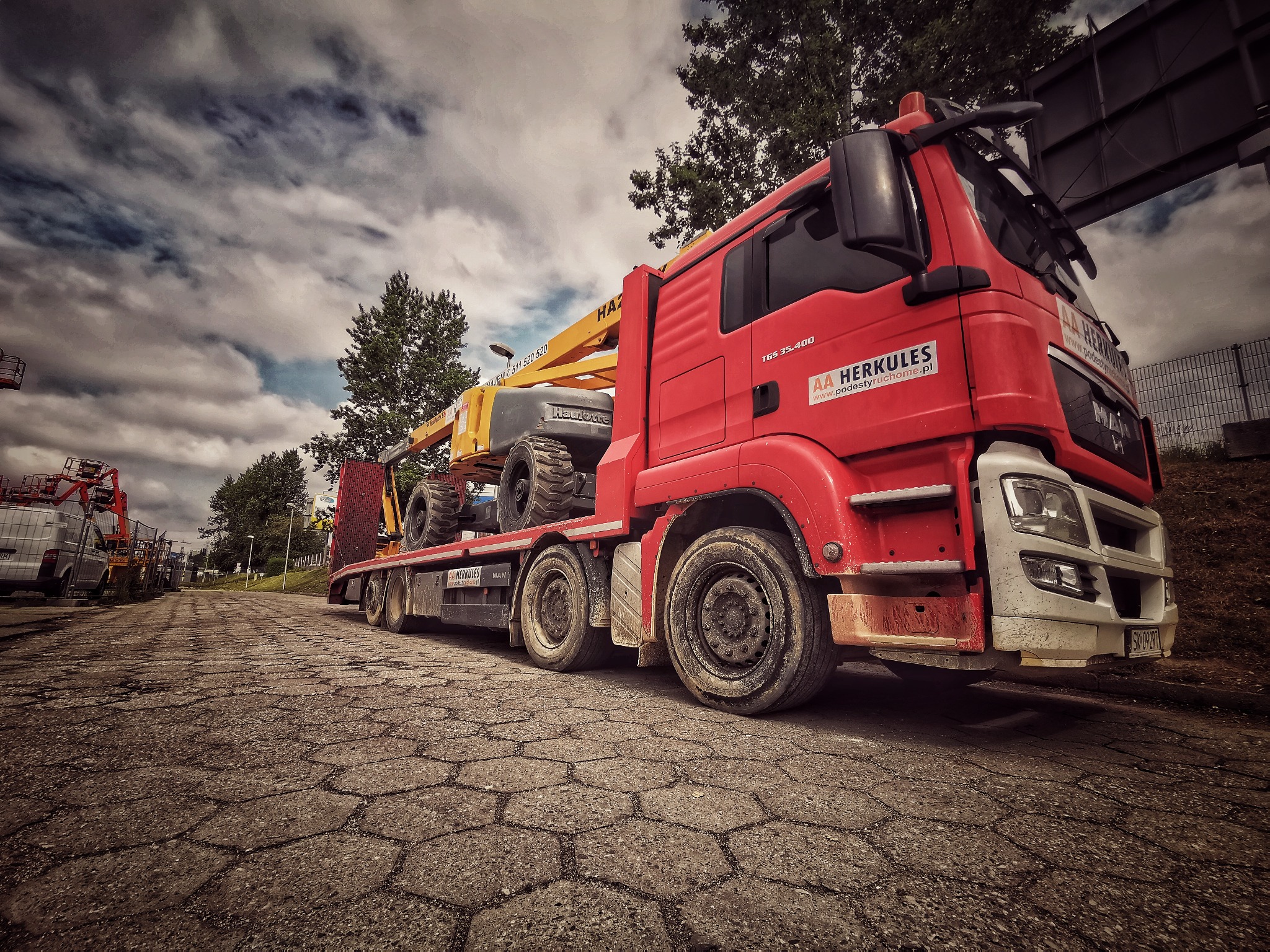 Czerwona laweta MAN z żółtą platformą roboczą Haulotte na naczepie, zaparkowana na wybrukowanej drodze pod pochmurnym niebem, logo firmy AA Herkules widoczne na kabinie.