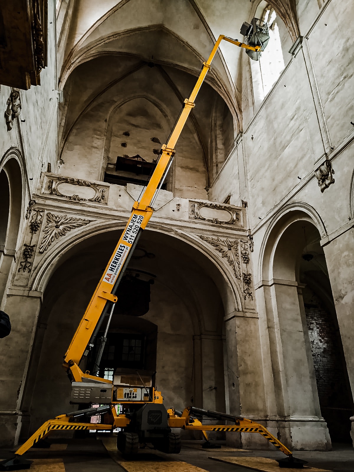 Żółty podnośnik koszowy wewnątrz zabytkowego kościoła z dekoracyjnymi łukami i filarami, platforma podnośnika przy oknie.