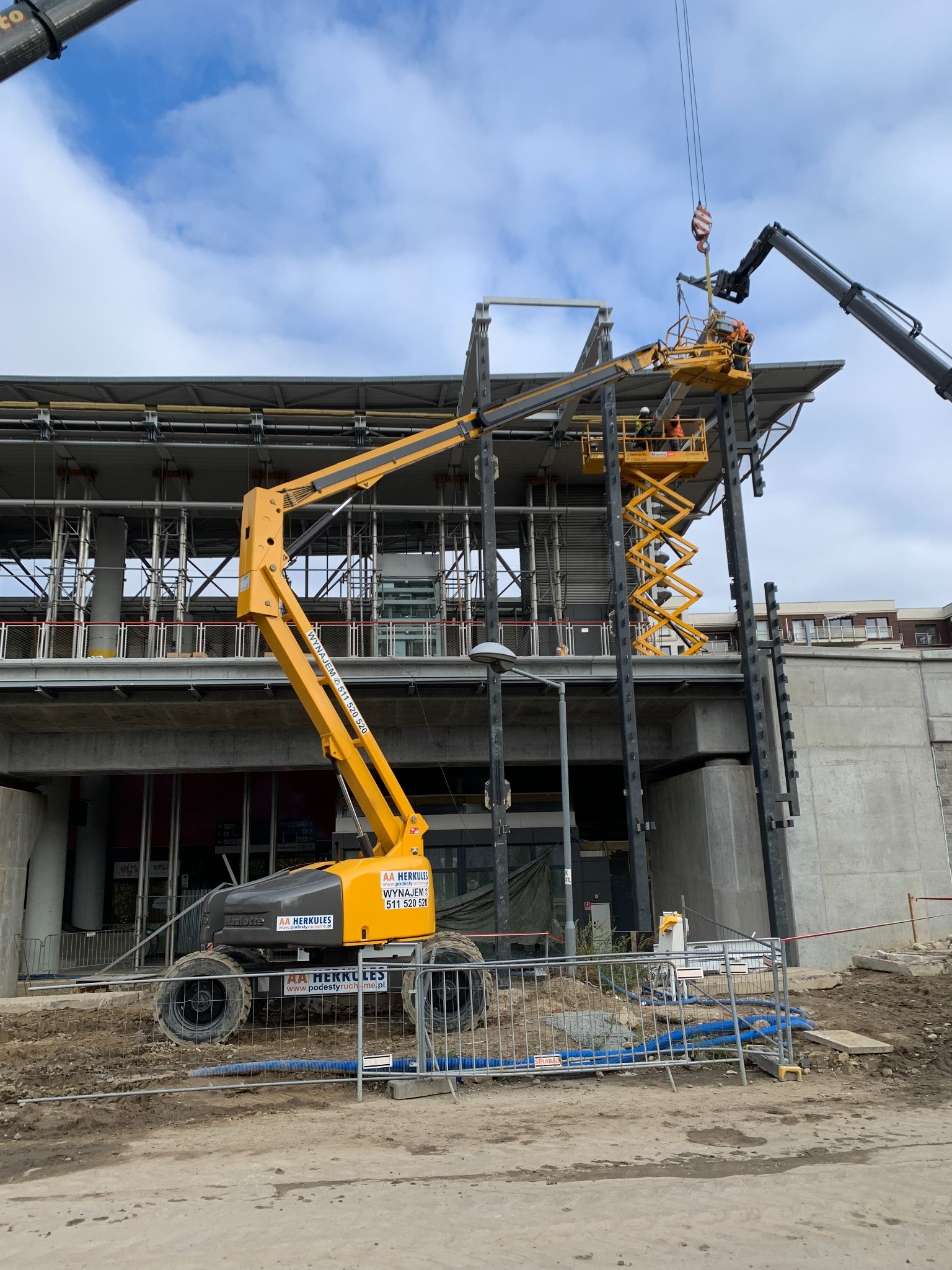 Żółty podnośnik koszowy i nożycowy na placu budowy, pracownicy w pomarańczowych kamizelkach na wysokościach, montaż konstrukcji stalowej, widoczny dźwig pomocniczy