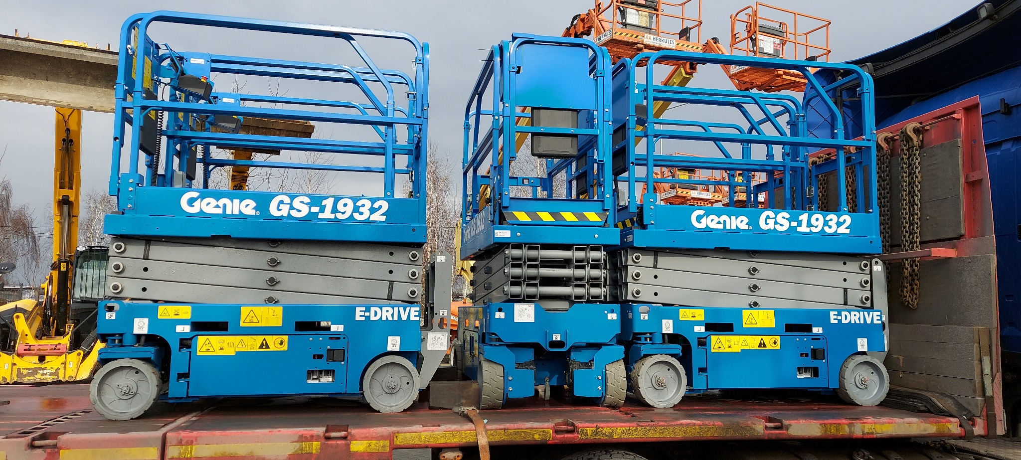 Dwie niebieskie platformy nożycowe Genie GS-1932 E-DRIVE załadowane na naczepę transportową, w tle widoczny podnośnik koszowy i żółta koparka.