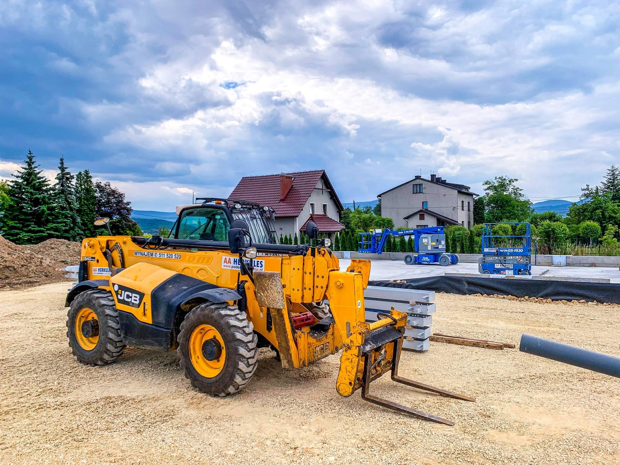 Żółty wózek widłowy teleskopowy JCB na placu budowy z przygotowanym fundamentem, w tle domy i pochmurne niebo.