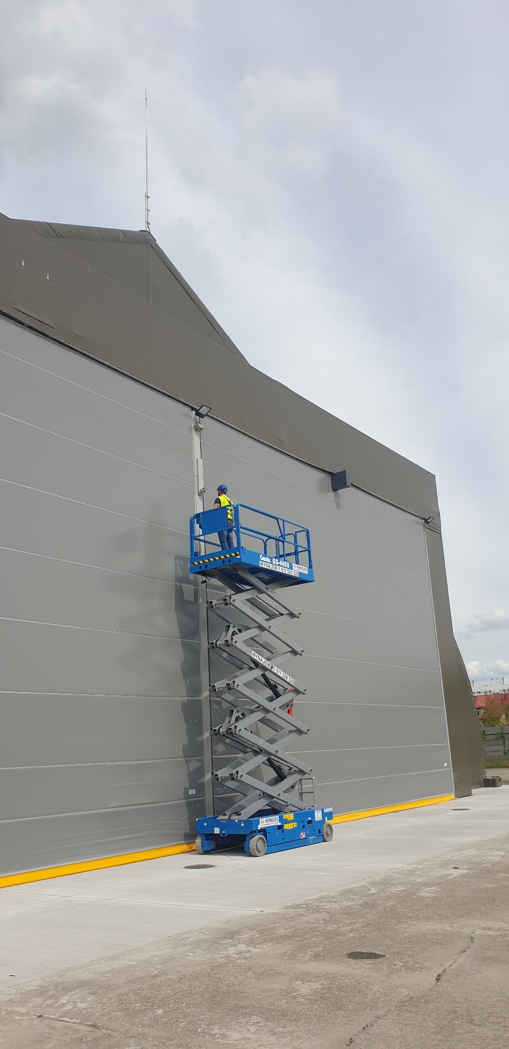 Pracownik w niebieskim podnośniku nożycowym, w kasku i kamizelce odblaskowej, dokonuje naprawy oświetlenia na szarej ścianie hali.