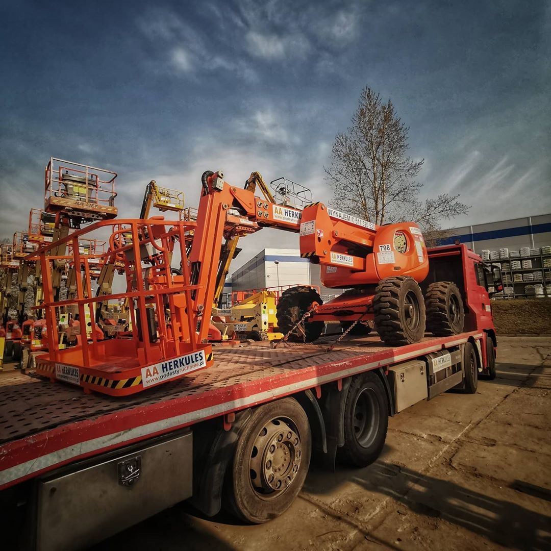 Pomarańczowa platforma robocza na lawecie ciężarówki w Katowicach, z widocznym logo firmy na koszu i korpusie, na tle magazynu i innych maszyn.