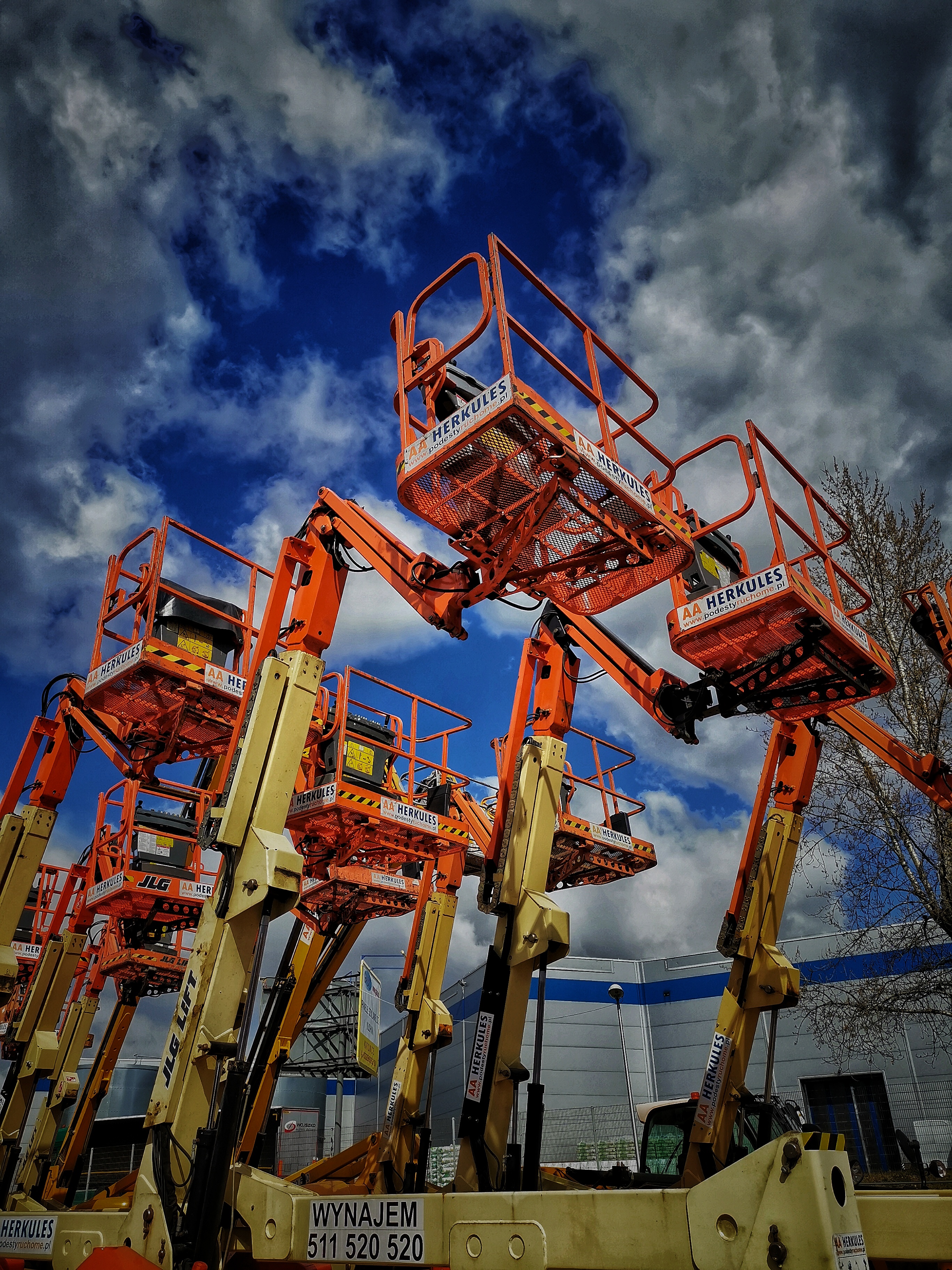 Rząd pomarańczowo-żółtych podnośników koszowych marek JLG i Herkules, ustawionych na placu, z błękitnym niebem i chmurami w tle.