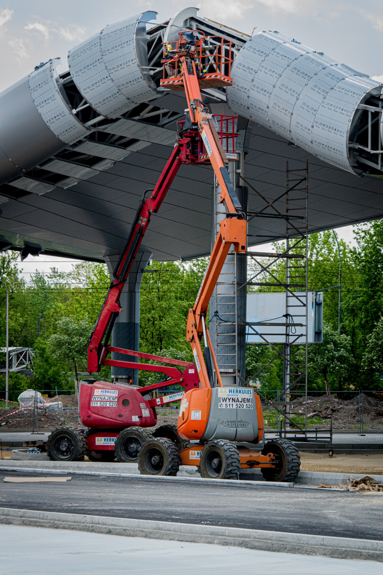 Dwie podnośniki koszowe, czerwona i pomarańczowa, pracują przy montażu metalowej konstrukcji z panelami na zewnątrz budynku. Operator w koszu pomarańczowego podnośnika. Widoczne oznaczenia firmy...
