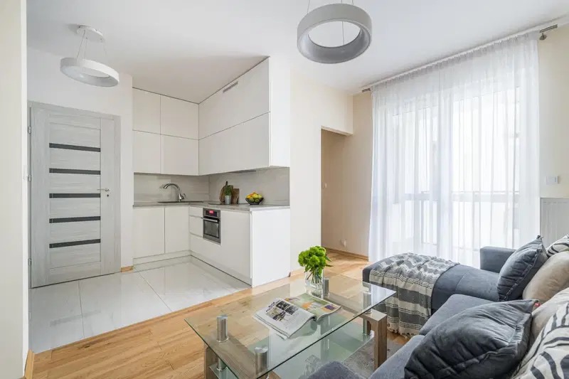 Jasne wnętrze mieszkania z aneksem kuchennym, białymi szafkami, szarą sofą z poduszkami w geometryczne wzory i szklanym stolikiem kawowym z gazetą.