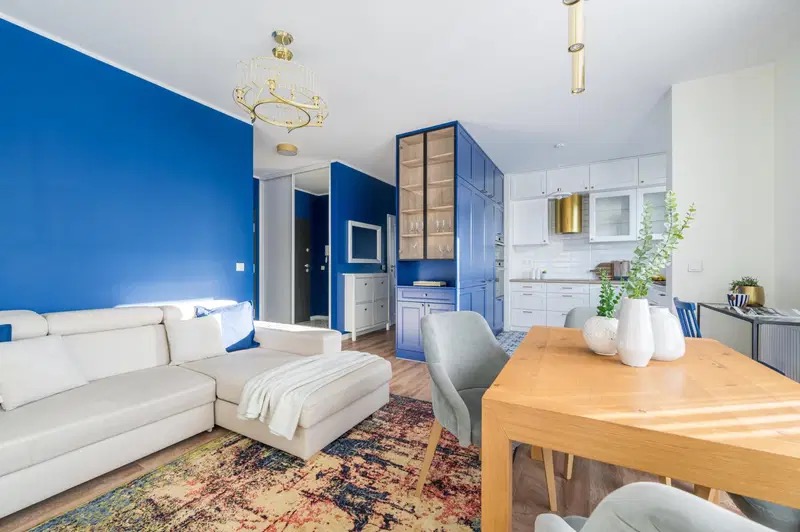 Jasny salon z niebieską ścianą, narożnikiem, drewnianym stołem i kuchnią w tle, oświetlony złotymi lampami.