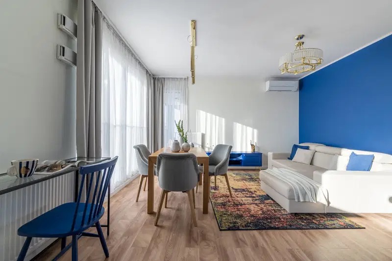 Jasne wnętrze mieszkania z drewnianą podłogą, stołem z szarymi krzesłami, białą sofą z niebieskimi poduszkami na tle niebieskiej ściany oraz kolorowym dywanem.