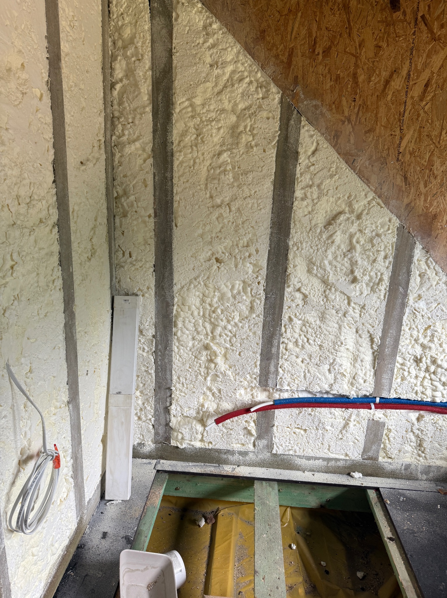 Ocieplony poddasze pianką PUR między krokwiami, widoczne instalacje i fragment podłogi. Biała pianka szczelnie wypełnia przestrzeń, szara krokiew.