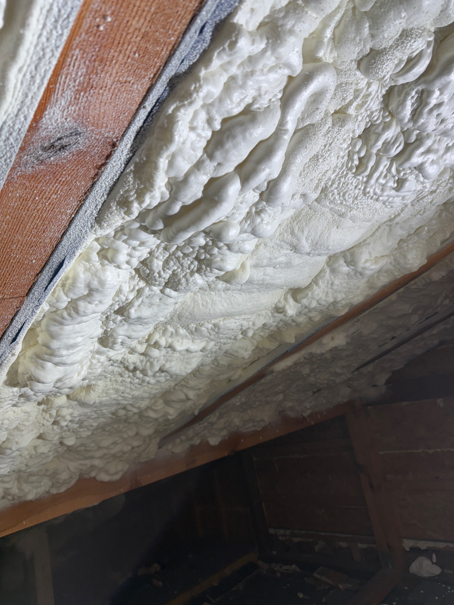 Ocieplenie poddasza pianką PUR, widoczne drewniane elementy konstrukcji. Biała, gęsta piana szczelnie wypełnia przestrzeń między krokwiami.