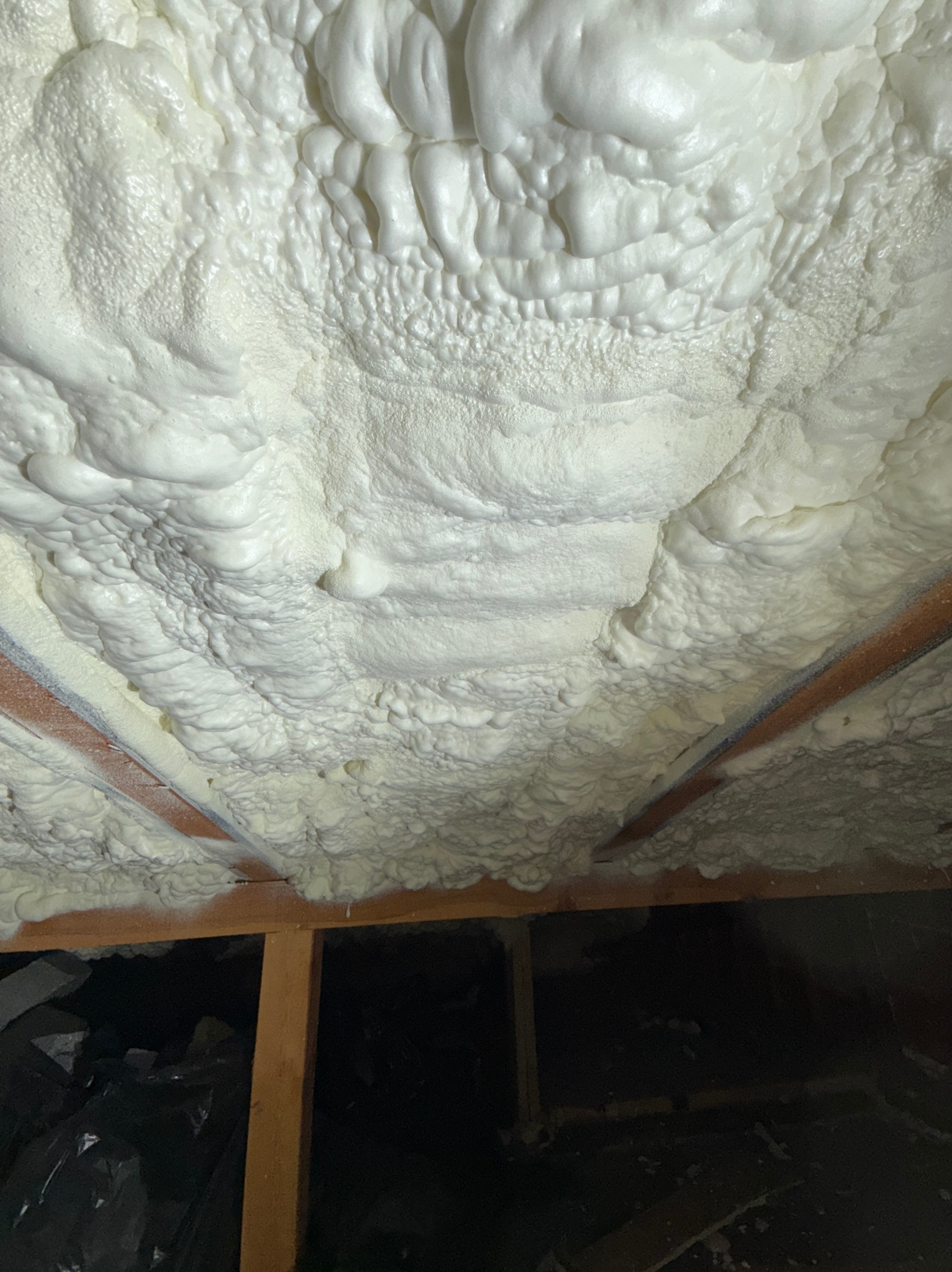 Szczelna izolacja poddasza pianką, widoczne drewniane elementy konstrukcji. Biała, gęsta struktura piany wypełnia przestrzeń między krokwiami.