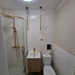 Nowoczesna łazienka z kabiną prysznicową ze złotymi elementami, umywalką z szafką w odcieniach drewna, toaletą i marmurowymi płytkami ściennymi z brązowymi żyłkami.