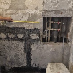 AUTORYZOWANY SERWIS DARIUSZ KASZOWSKI - Łazienka w trakcie demontażu, widoczne uszkodzone ściany, stare rury, toaleta zarysowana czerwoną kredką i narzędzia do skuwania płytek na podłodze, osoba mierzy ścianę taśmą mierniczą.