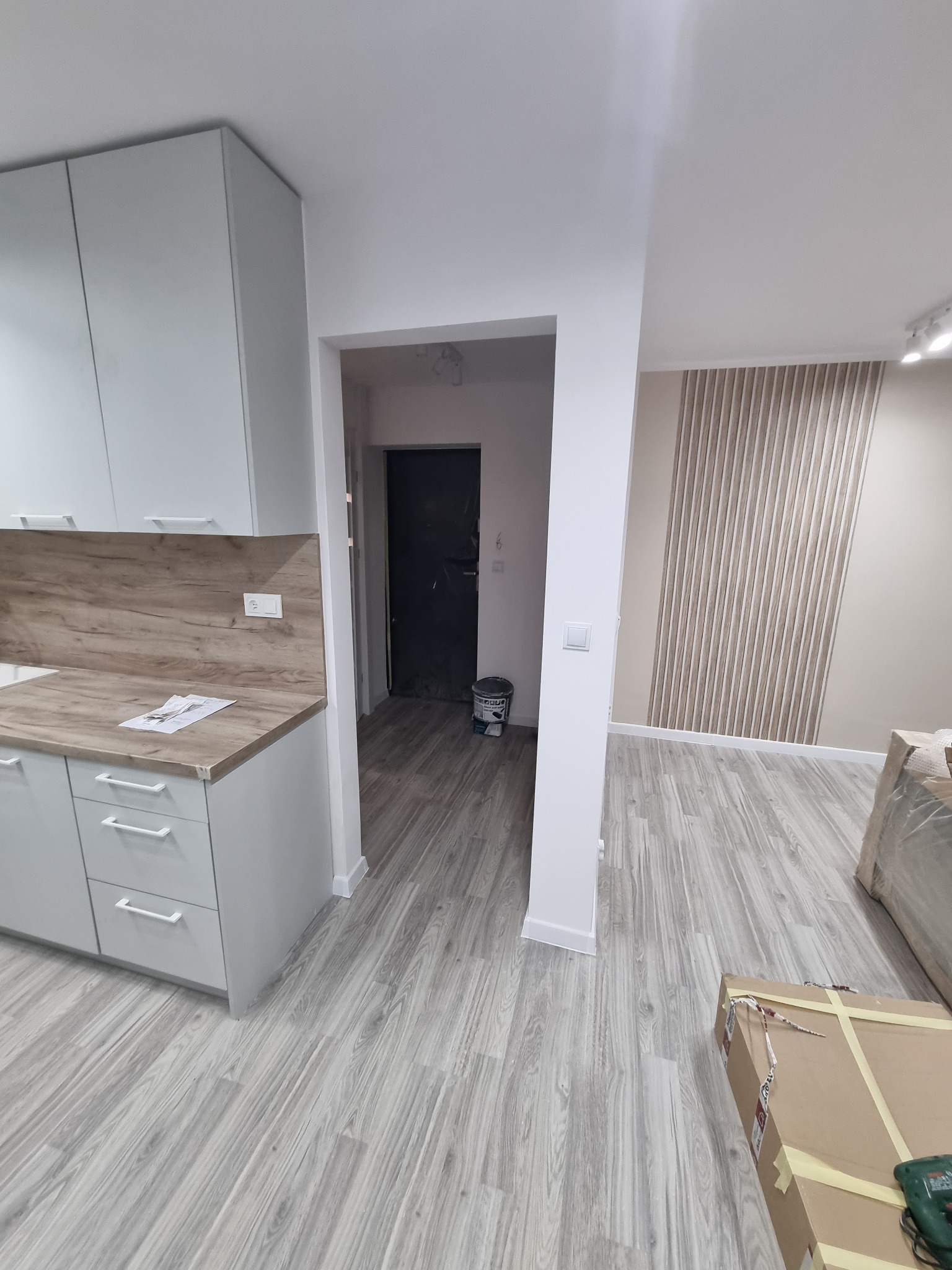 Nowoczesna kuchnia z szarymi szafkami i drewnianym blatem, widok na korytarz z ciemnymi drzwiami, panele podłogowe imitujące drewno, dekoracyjna ściana z pionowymi panelami, kartonowe pudła w rogu...