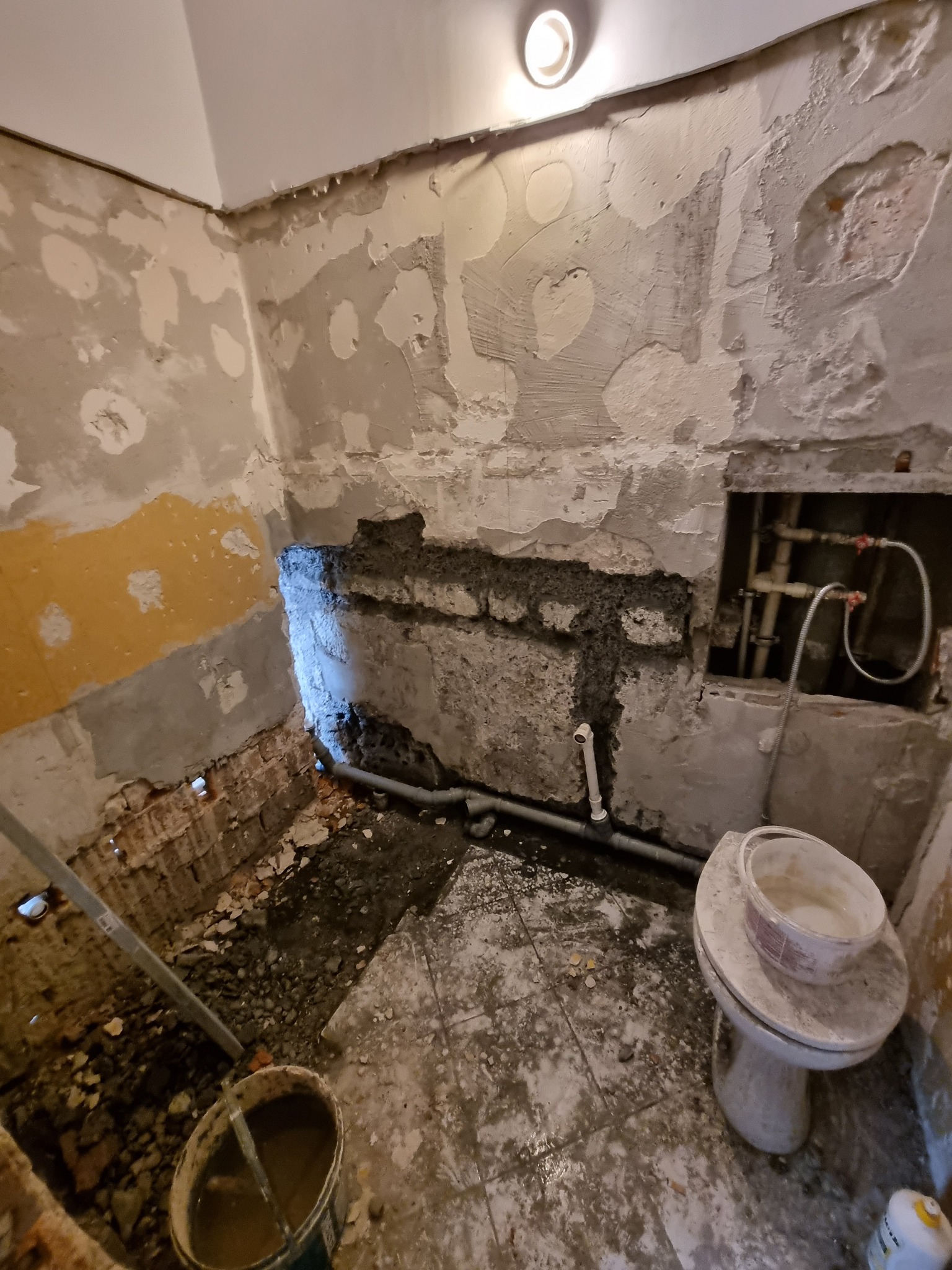 Łazienka w trakcie demontażu: odsłonięte rury, resztki tynku na ścianach, gruz na podłodze, wiadro z zaprawą i sedes z resztkami farby.