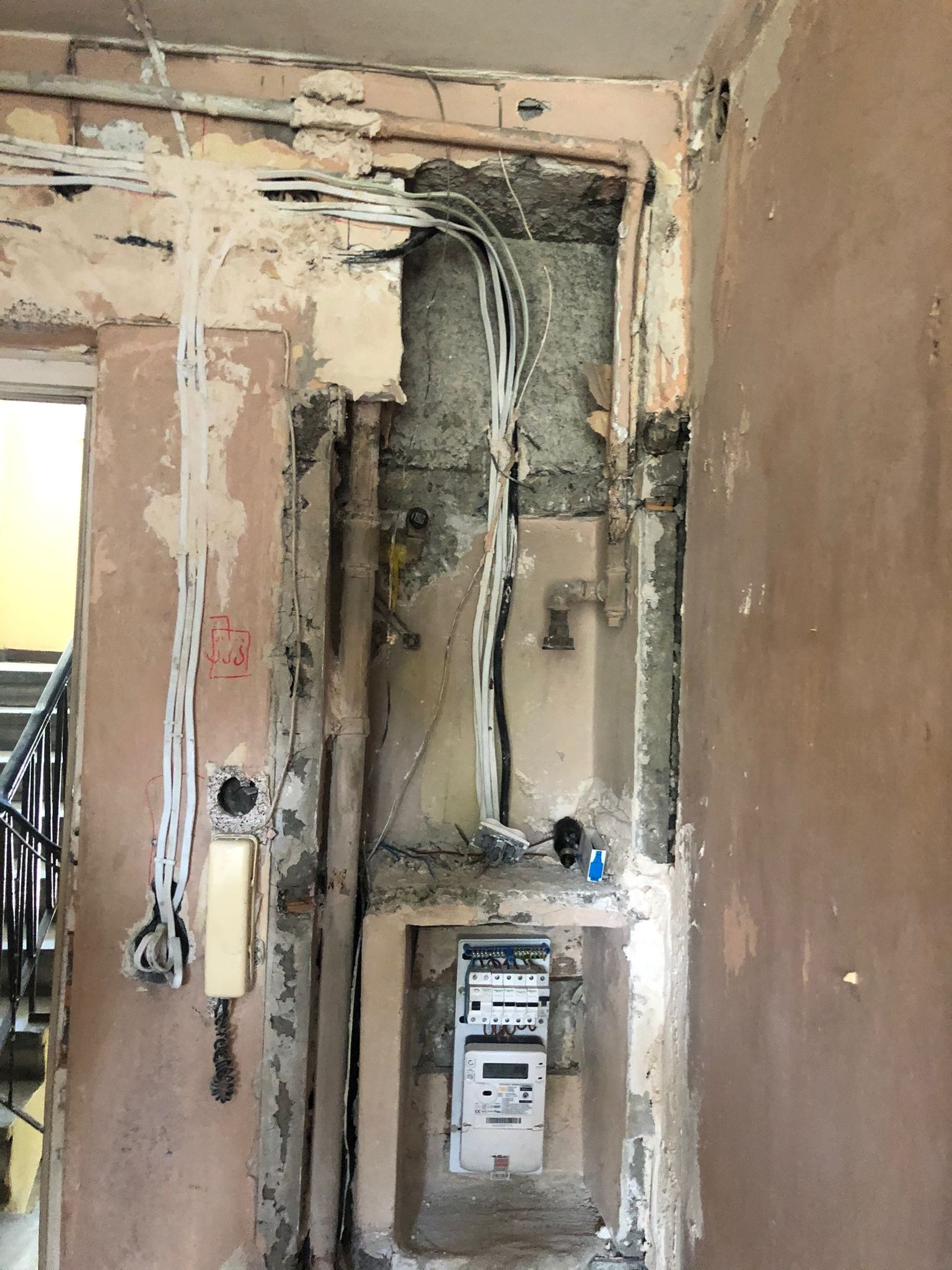 Korytarz w trakcie remontu: odsłonięte przewody elektryczne, rury, licznik energii elektrycznej w niszy, stara instalacja domofonowa obok schodów.