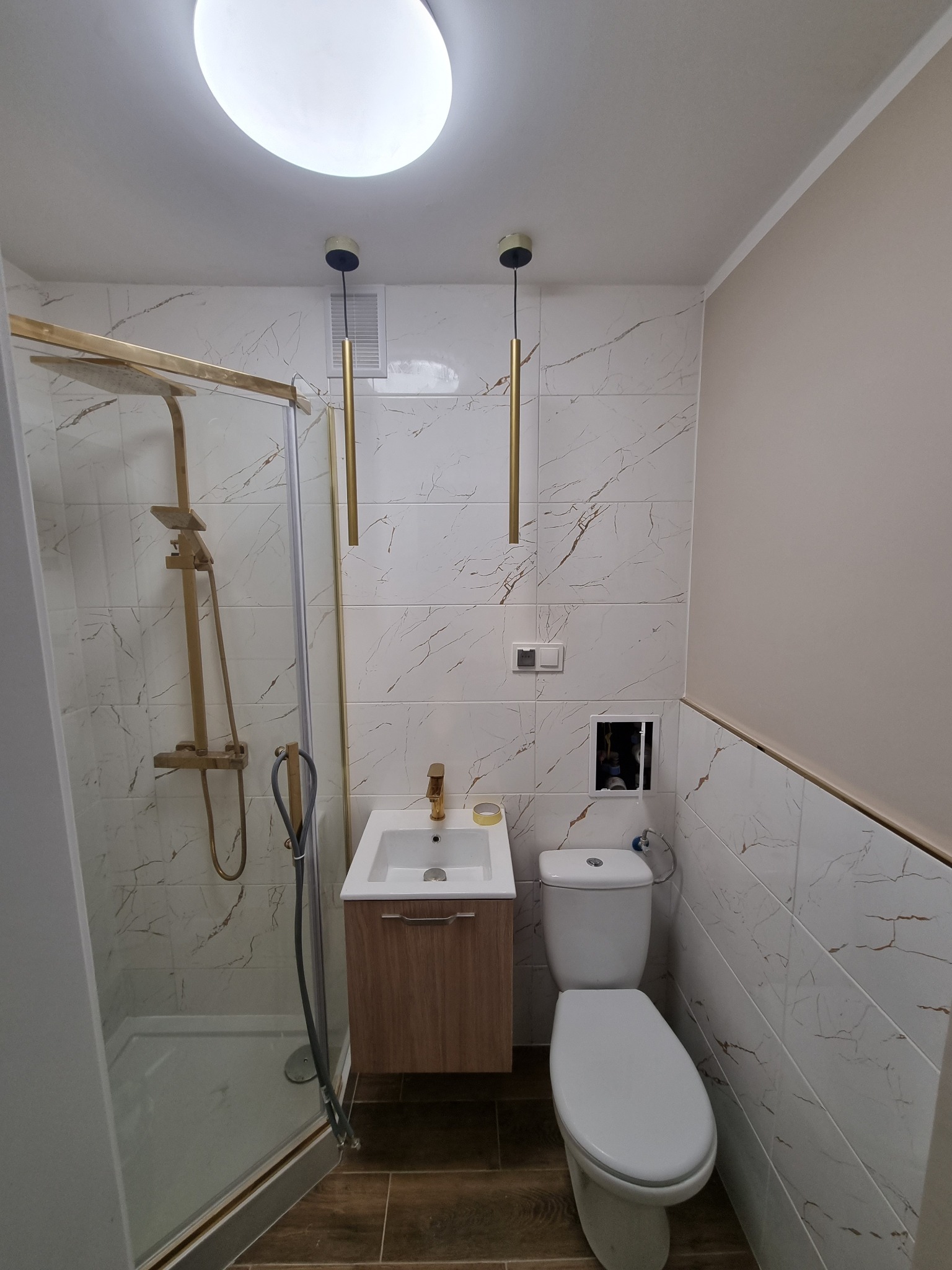 Nowoczesna łazienka z kabiną prysznicową ze złotymi elementami, umywalką z szafką w odcieniach drewna, toaletą i marmurowymi płytkami ściennymi z brązowymi żyłkami.