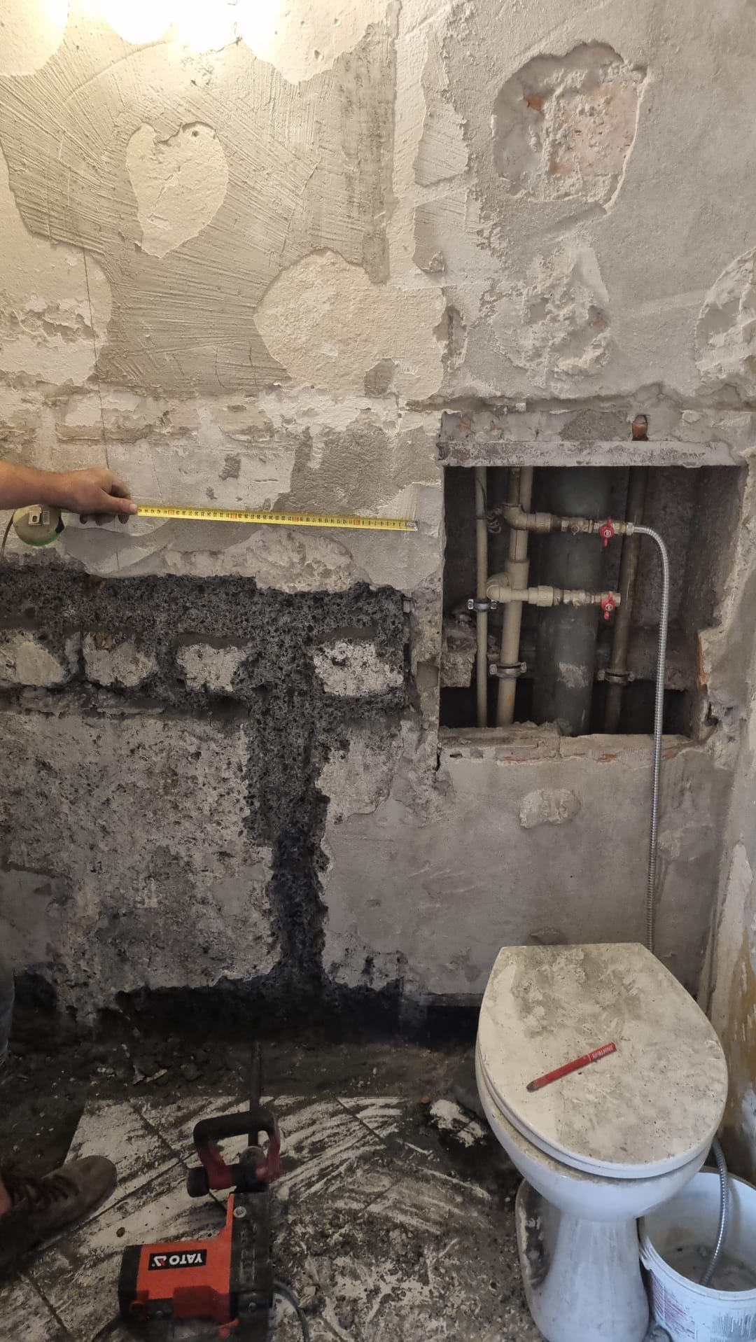 Łazienka w trakcie demontażu, widoczne uszkodzone ściany, stare rury, toaleta zarysowana czerwoną kredką i narzędzia do skuwania płytek na podłodze, osoba mierzy ścianę taśmą mierniczą.