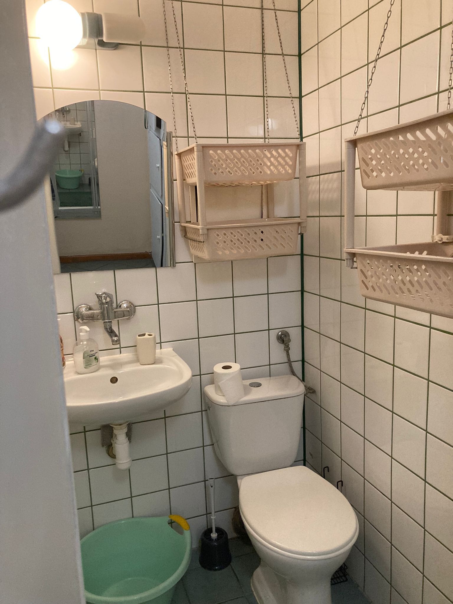 Standardowa łazienka w Koszalinie z białymi kafelkami, umywalką, toaletą i plastikowymi koszykami wiszącymi na łańcuchach, widoczna z perspektywy wejścia.
