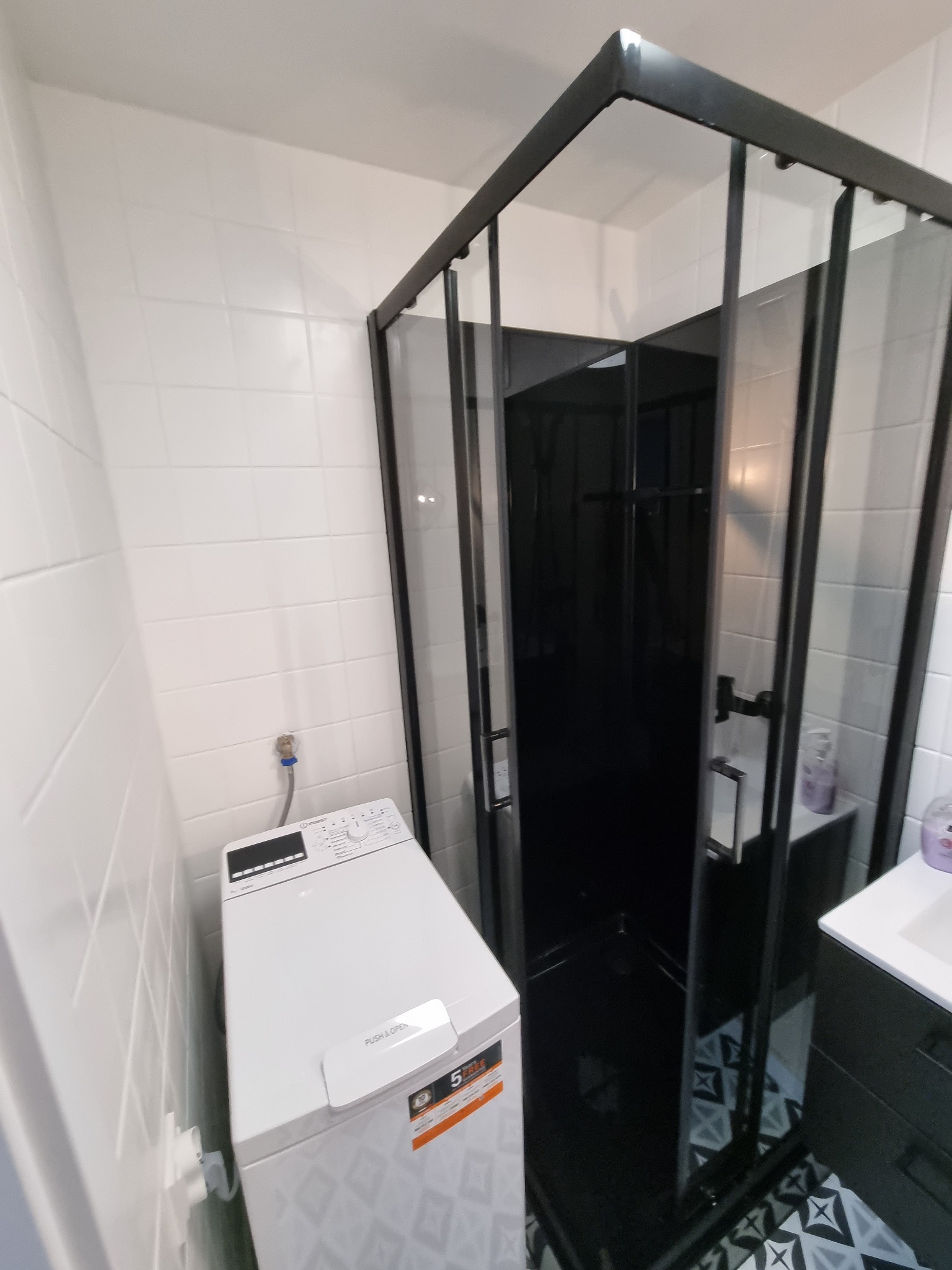 Nowoczesna łazienka z kabiną prysznicową typu walk-in z czarnymi profilami, białą pralką z przodu i czarno-białą mozaiką na podłodze.