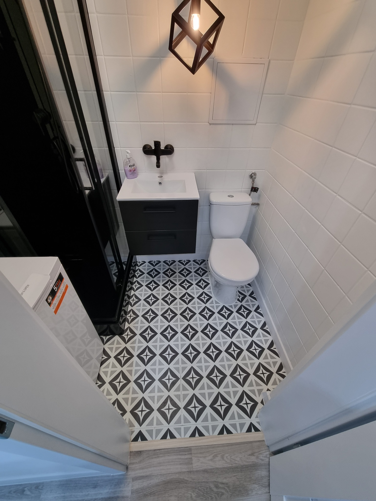 Nowoczesna łazienka z białą armaturą, czarną szafką pod umywalką i kabiną prysznicową typu walk-in z czarnymi ramami, wzorzysta podłoga z motywem gwiazdy, oświetlenie punktowe.