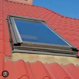 Okno dachowe z szarą ramą zamontowane na czerwonym dachu pokrytym dachówką ceramiczną; widoczna membrana uszczelniająca wokół okna.