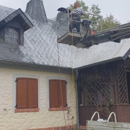 Dekarstwo-Blacharstwo - Pracownik na podnośniku koszowym przy dachu domu z szarej dachówki łupkowej; widoczne kominy z daszkami, brązowe okiennice i żółte kwiaty w ogrodzie.