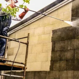 Pracownik w kombinezonie i rękawicach ochronnych, stojący na rusztowaniu, czyści ścianę budynku za pomocą myjki ciśnieniowej, widoczna różnica między czystą a zabrudzoną powierzchnią.