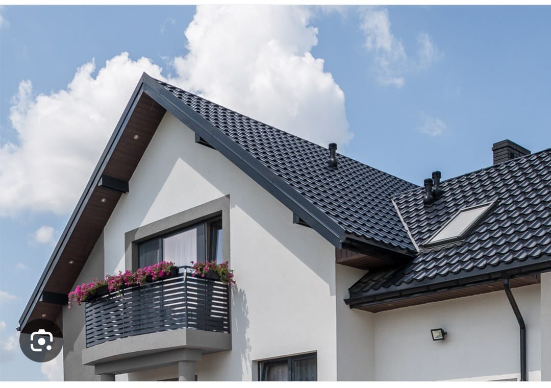 Dom jednorodzinny z ciemną, metalową dachówką, balkonem z kwiatami i widocznymi kominami wentylacyjnymi.