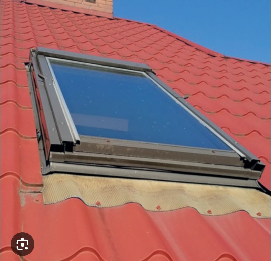 Okno dachowe z szarą ramą zamontowane na czerwonym dachu pokrytym dachówką ceramiczną; widoczna membrana uszczelniająca wokół okna.
