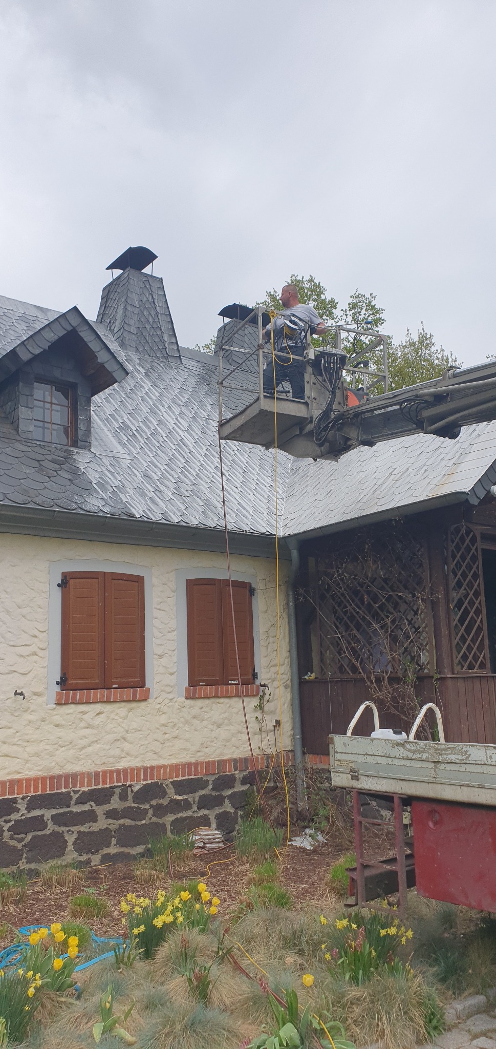 Pracownik na podnośniku koszowym przy dachu domu z szarej dachówki łupkowej; widoczne kominy z daszkami, brązowe okiennice i żółte kwiaty w ogrodzie.