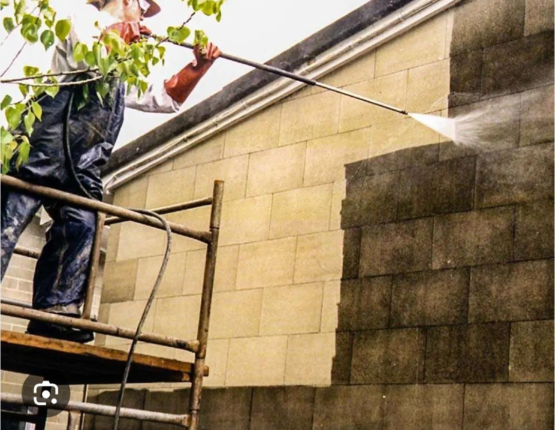 Pracownik w kombinezonie i rękawicach ochronnych, stojący na rusztowaniu, czyści ścianę budynku za pomocą myjki ciśnieniowej, widoczna różnica między czystą a zabrudzoną powierzchnią.