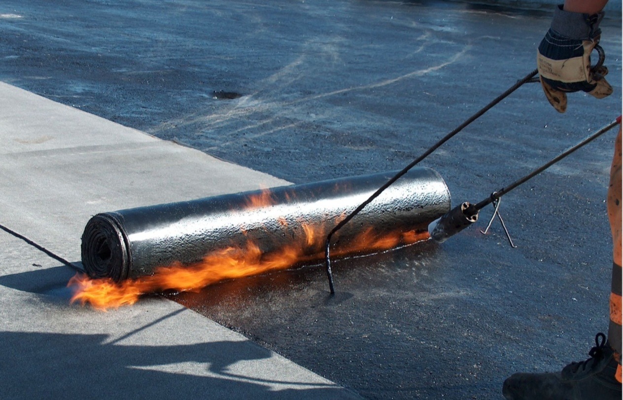 Pracownik w rękawicach spawa papę termozgrzewalną na płaskim dachu, płomienie ognia widoczne spod rolki.