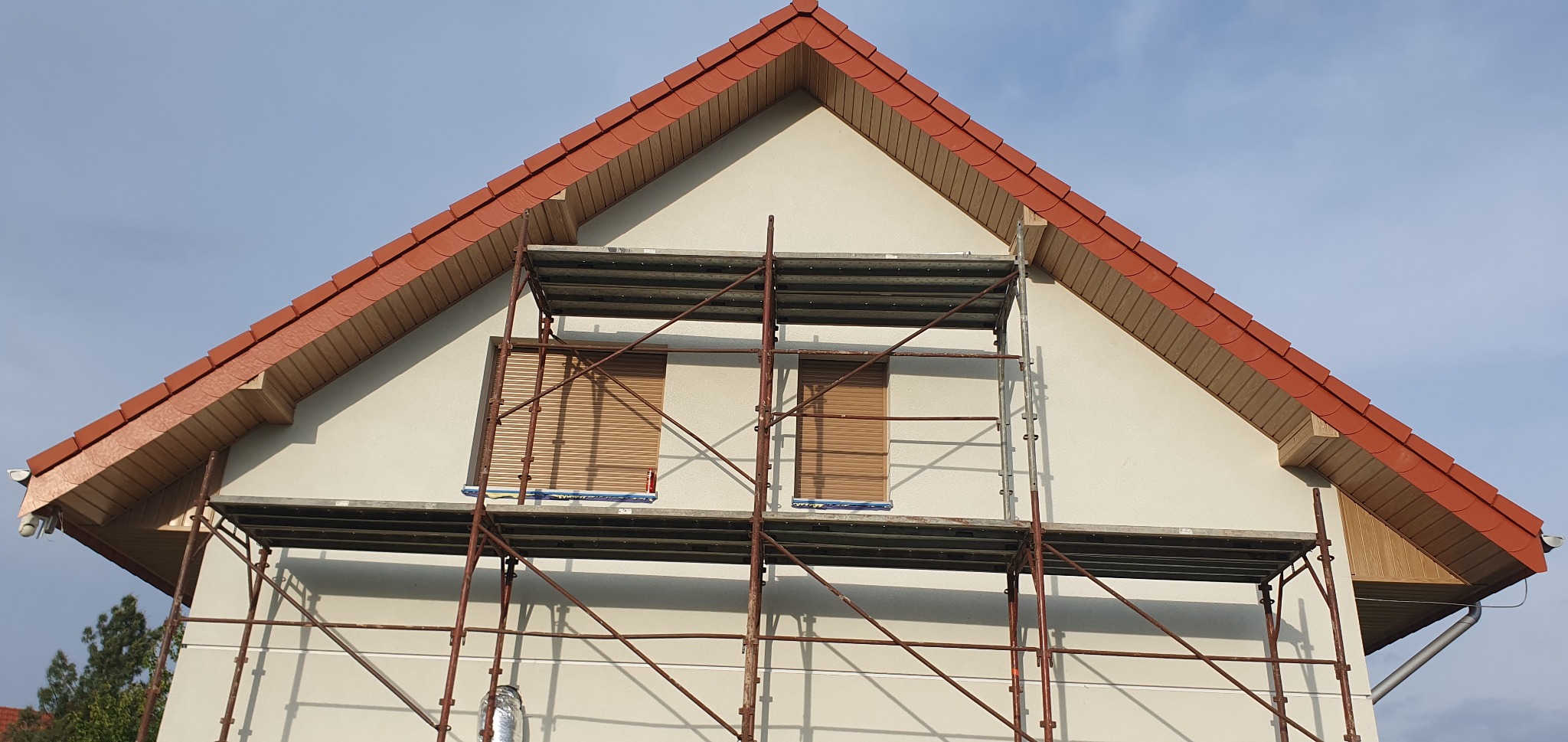 Dom jednorodzinny w trakcie prac na wysokości, rusztowanie przy oknie, widoczny fragment dachu z czerwoną dachówką. Ściana budynku w jasnym kolorze.