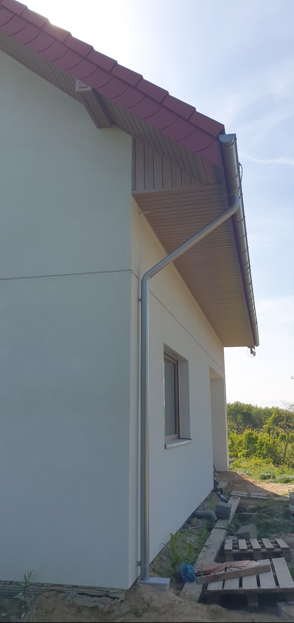 Nowa, metalowa rynna pionowa na elewacji jasnego domu jednorodzinnego z dachem pokrytym czerwoną dachówką. Widoczne okno i podbitka dachowa.